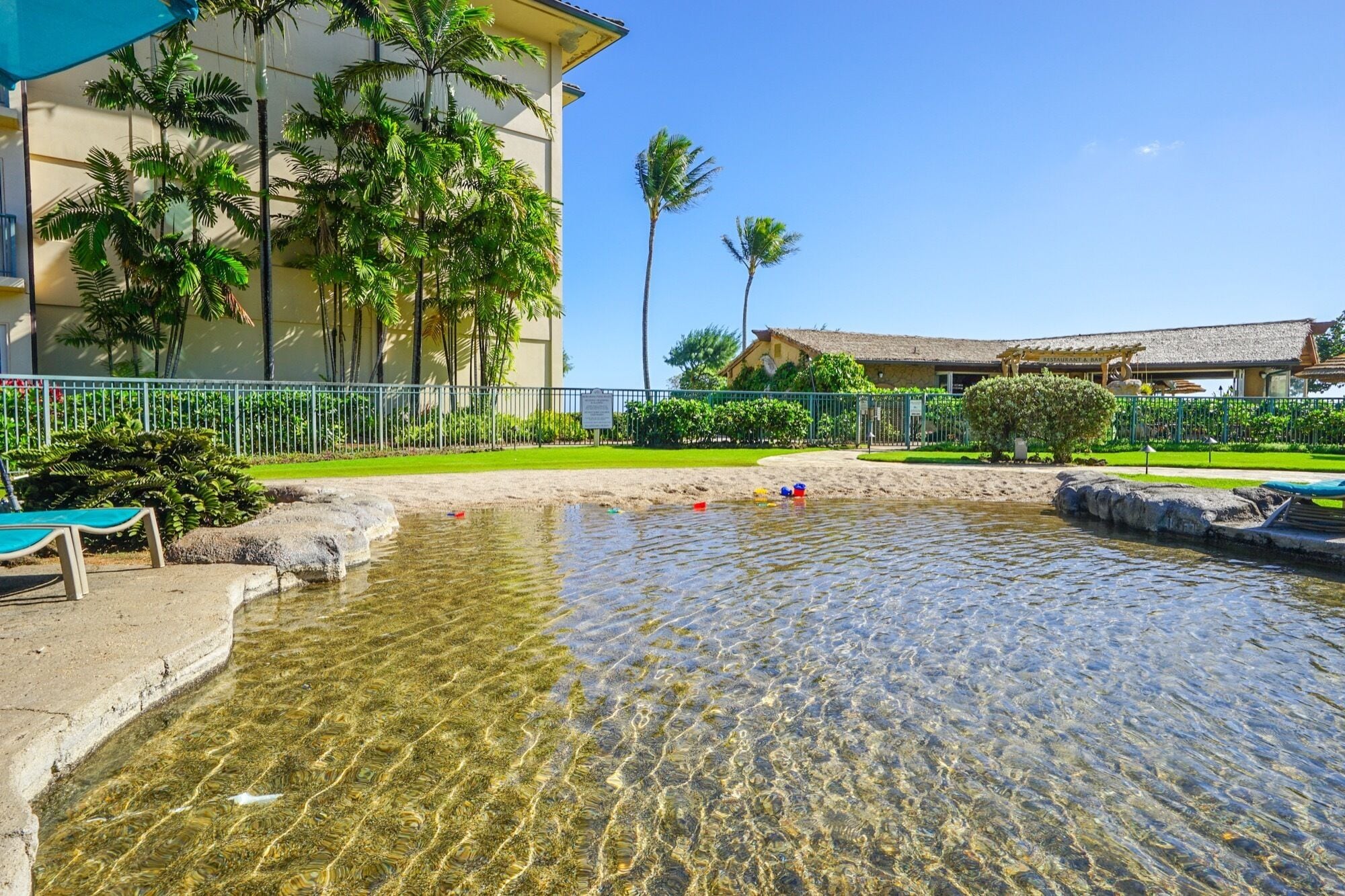 Una piscina cristalina con zona de arena y palmeras, rodeada por una valla y un edificio con letrero de restaurante.