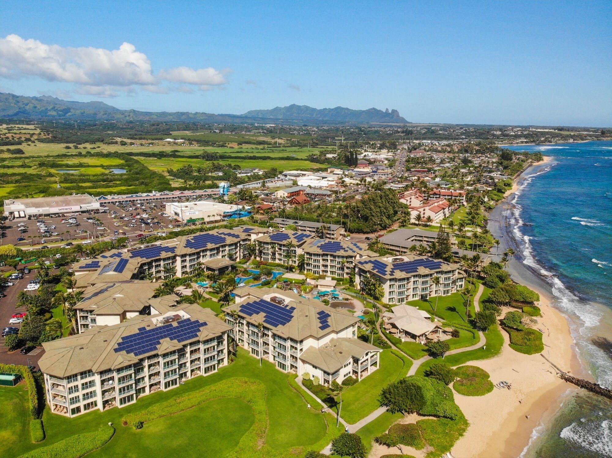 Un resort con varias construcciones que incluyen paneles solares, una playa y montañas al fondo.
