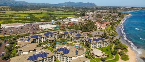 Un resort con varias construcciones que incluyen paneles solares, una playa y montañas al fondo.