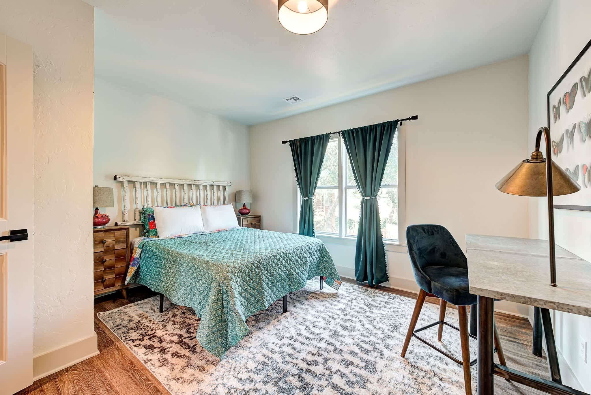 A bedroom with a bed, a desk, a chair, a radiator, and a window with curtains.