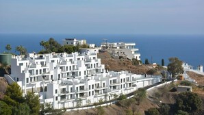 A coastal residential area with modern white buildings, a clear view of the sea, and a hilly landscape.