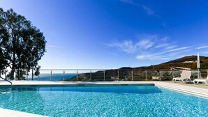 A swimming pool with clear blue water, a metal railing, and a view of the ocean and hills in the background.
