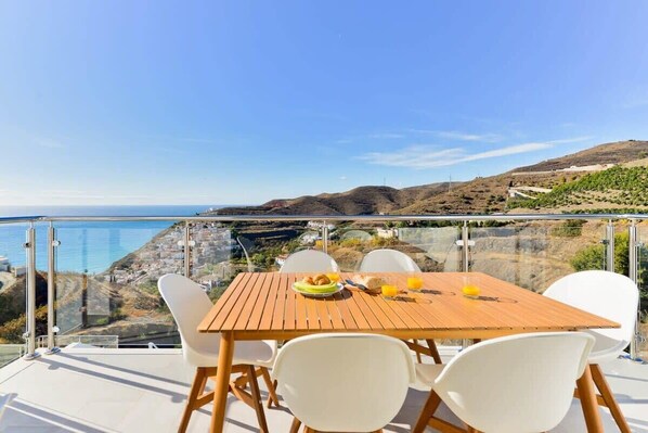 Une table avec des chaises et une assiette de nourriture sur un balcon donnant sur la mer et les collines.