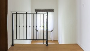 A hallway with wooden flooring, a black metal railing, a ceiling light fixture, and a window with sheer curtains.