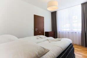 Un lit bien fait dans une chambre d’hôtel, avec une porte en bois, une table de chevet et une fenêtre avec des rideaux.