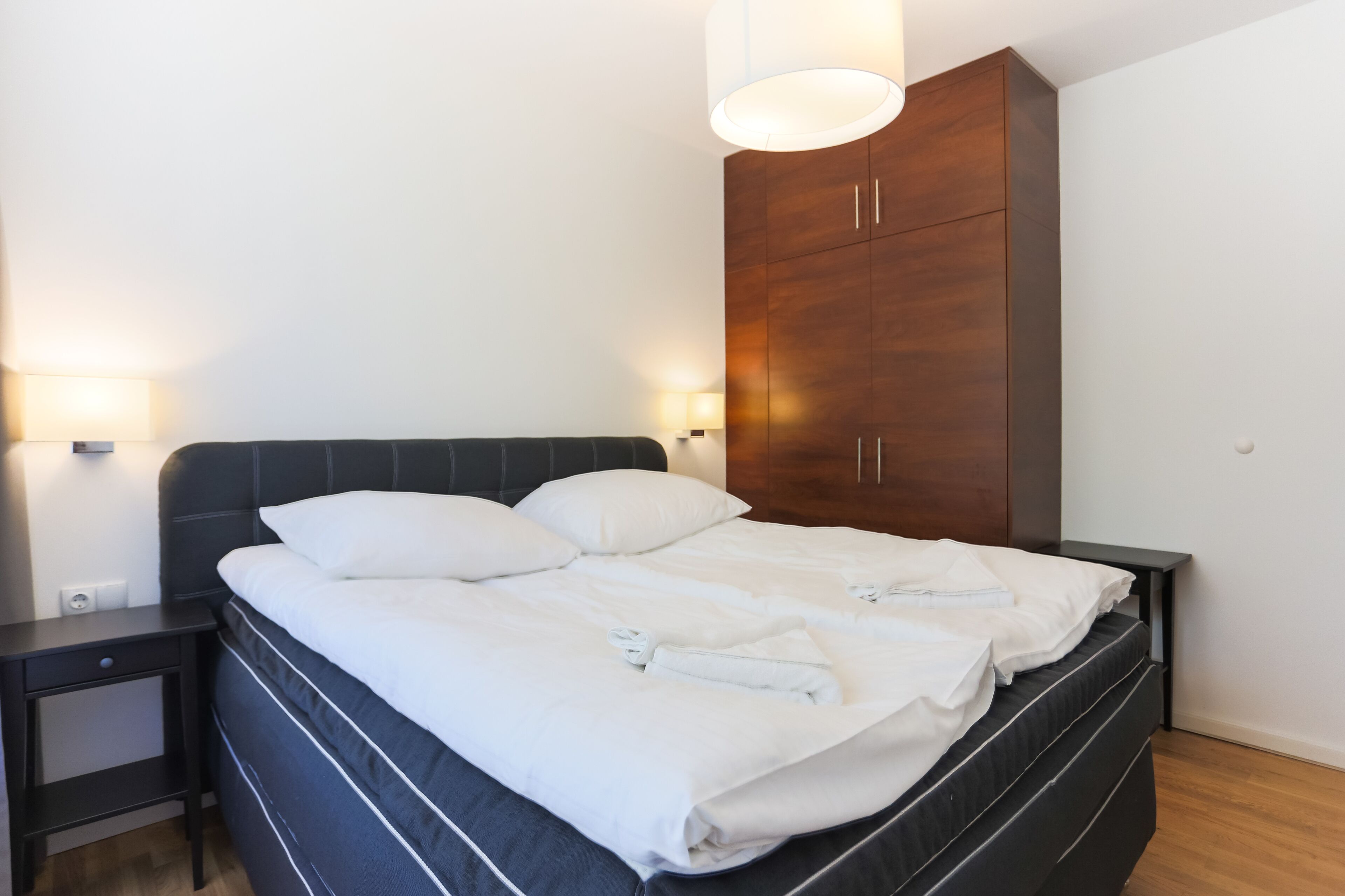Una cama doble con ropa blanca de cama bien tendida, un cabecero oscuro y un armario de madera con dos puertas.