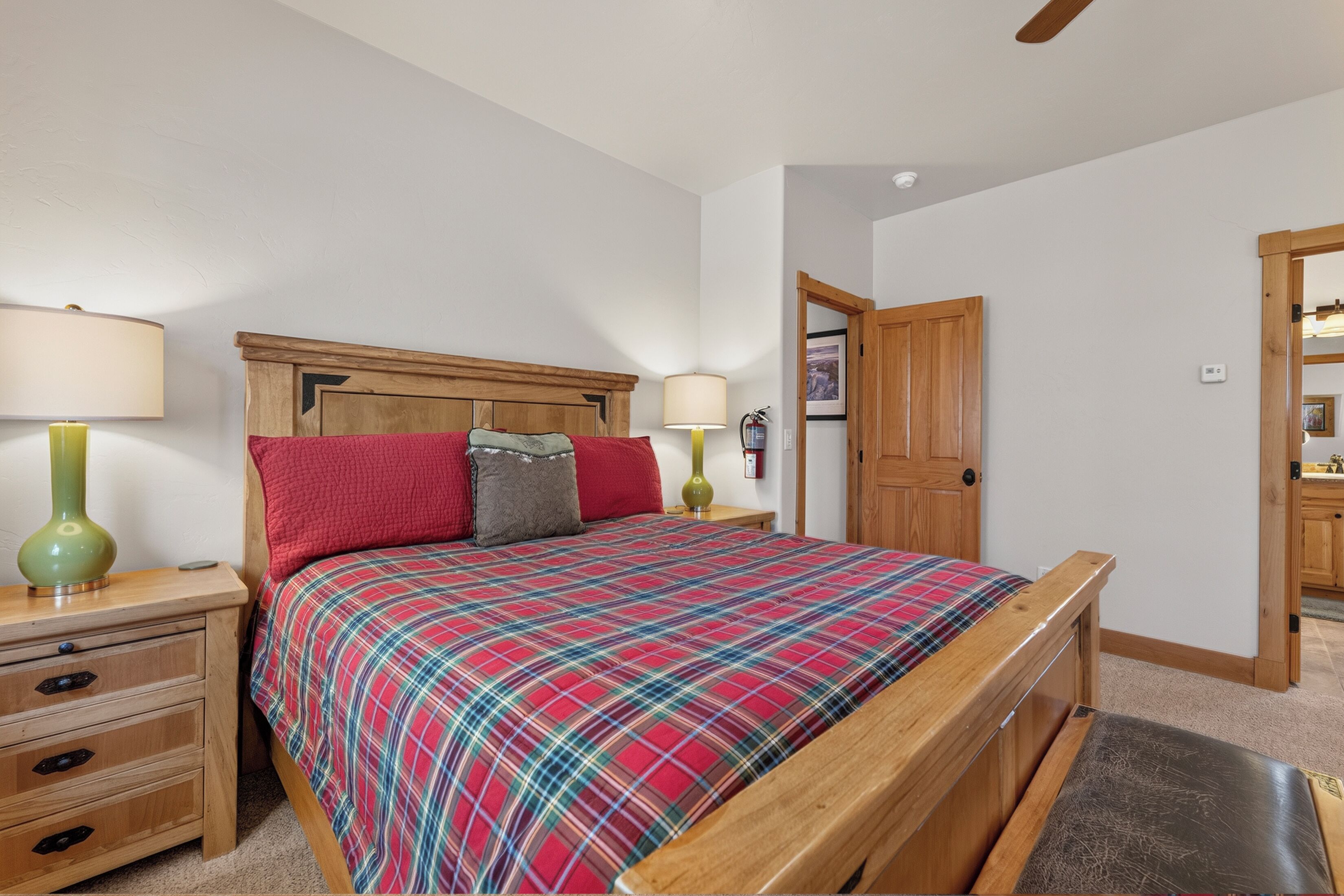 A bedroom with a wooden bed frame, a plaid bedspread, two bedside tables with lamps, a wooden nightstand, and a door leading to another room.