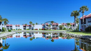 A resort with a central water feature reflecting buildings, palm trees, and a clear blue sky.