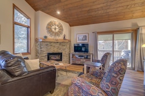 A cozy living room with a stone fireplace, leather sofa, patterned armchair, and a large clock on the wall.