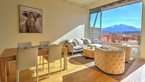 A modern living room with a dining area, large windows with a mountain view, and a painting of a cow on the wall.