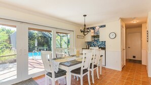 Comedor con una mesa preparada para cuatro, sillas blancas, una araña y vistas a una piscina a través de puertas de cristal.