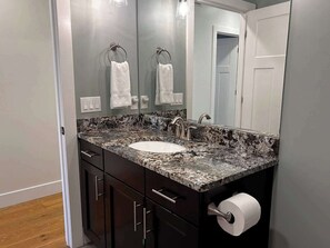 A bathroom with a granite countertop sink, dark cabinetry, a mirror, and a toilet paper holder.