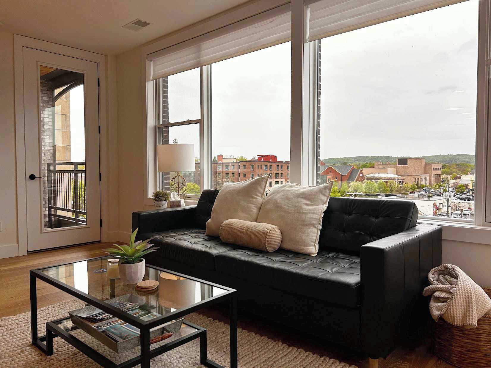 A modern living room with a black leather sofa, a glass coffee table, and a view of a cityscape through large windows.