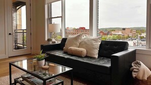 Un salon moderne avec un canapé en cuir noir, une table basse en verre et une vue sur le paysage urbain grâce à de grandes fenêtres.