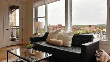 A modern living room with a black leather sofa, a glass coffee table, and a view of a cityscape through large windows.