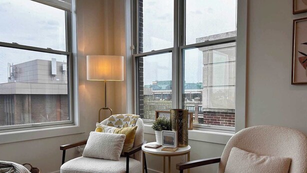 Une pièce chaleureuse avec deux fauteuils, une lampe sur pied et une petite table agrémentée d’objets décoratifs. De grandes fenêtres offrent une vue sur le paysage urbain.
