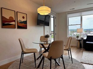 A modern living room with a glass dining table, four chairs, a sofa, a TV, and artwork on the wall.