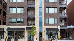 Un immeuble à plusieurs étages doté d’un élément vertical en verre au centre, de nombreux balcons et de boutiques au niveau du rez-de-chaussée.