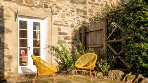 A stone building with a white door, two wicker chairs, and a wooden fence. - Charming Cardoon Cottage in Beautiful Village (Crieff)
