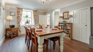A dining room with a wooden table set for four, surrounded by chairs. There is a bookshelf filled with books to the right. - Charming Cardoon Cottage in Beautiful Village (Crieff)