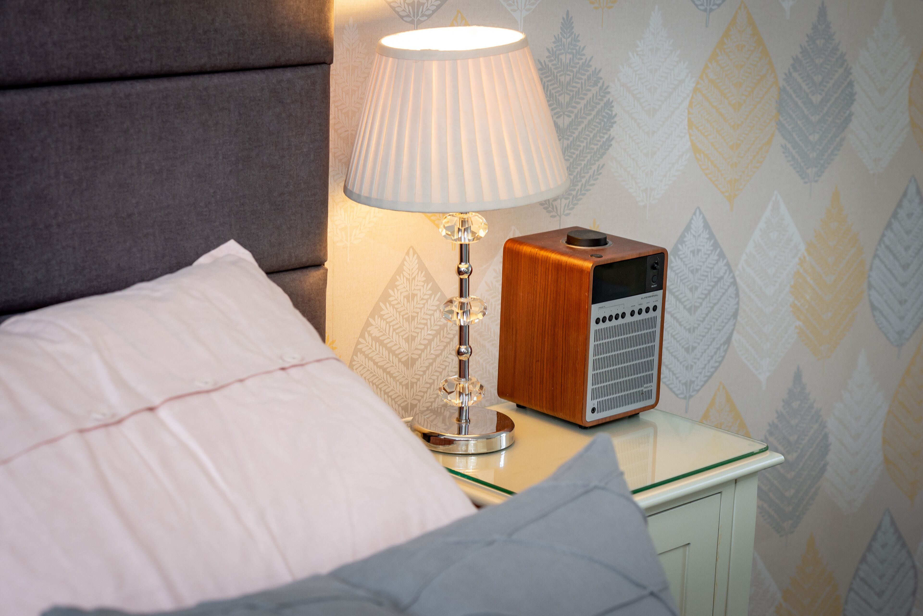 Ein ordentlich bezogenes Bett mit einem weißen Kopfkissen, einer Nachttischlampe und einem Nachttisch aus Holz mit einem Radio.