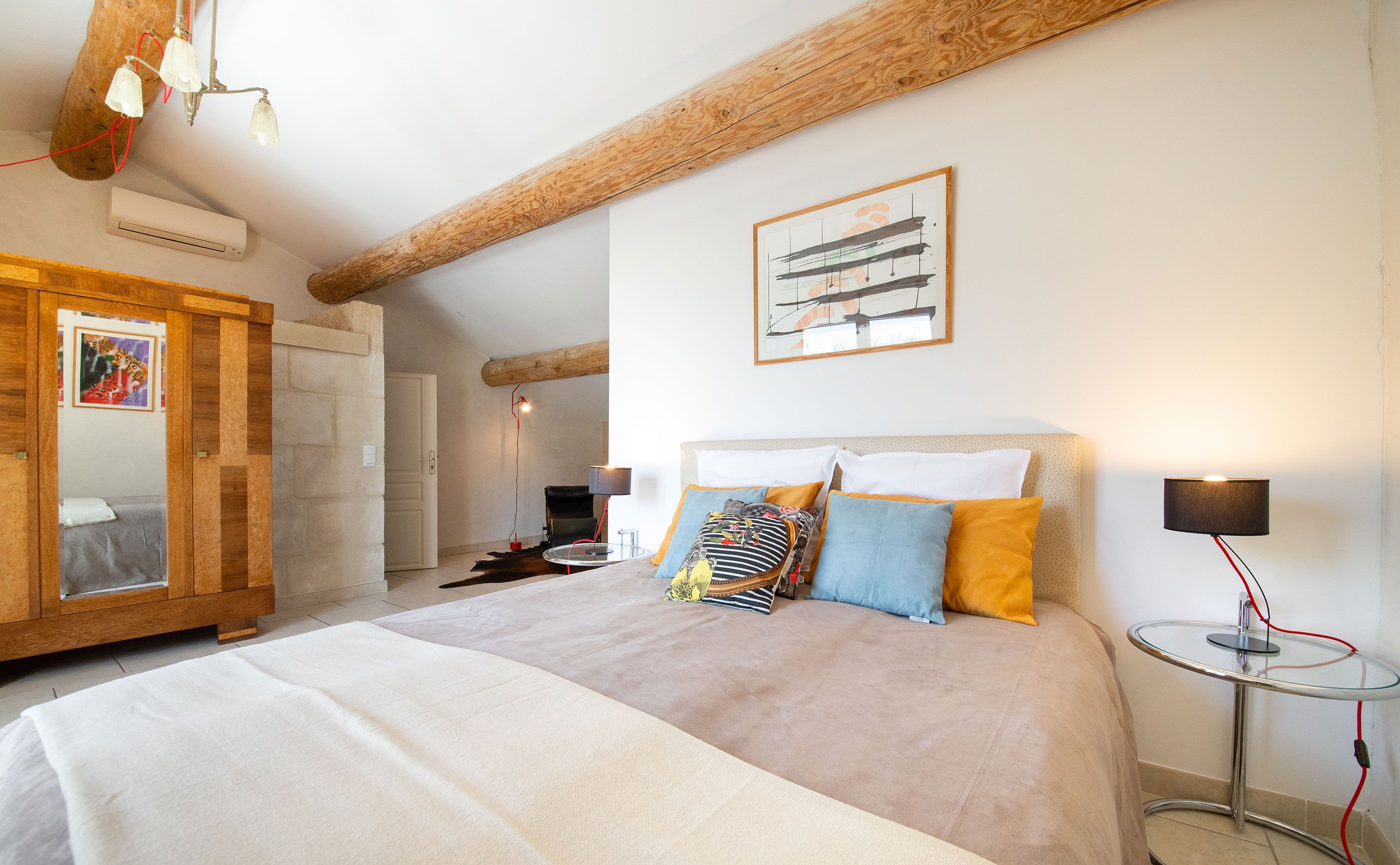 Une chambre avec un grand lit, des poutres apparentes, une petite table avec une lampe et un tableau encadré au mur.
