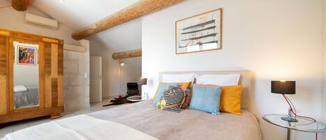 Een slaapkamer met een groot bed, houten balken, een klein tafeltje met een lamp en een ingelijste foto aan de muur.