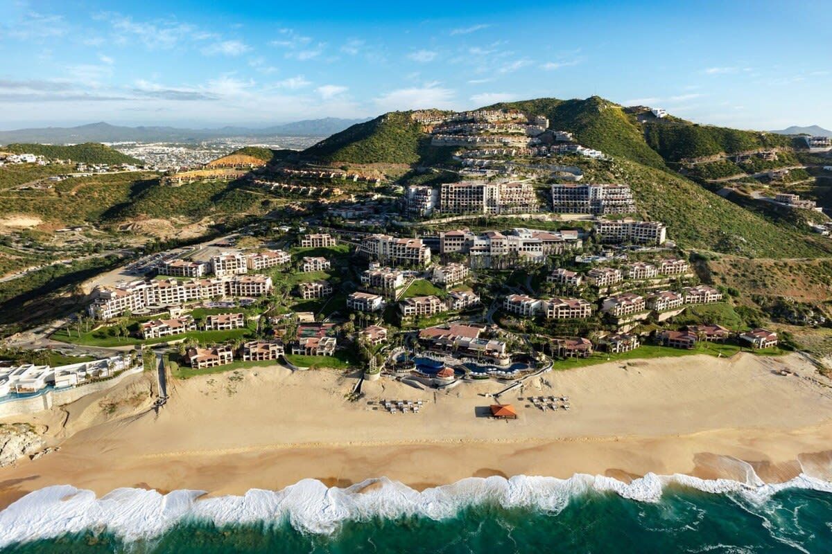 Un complejo turístico en la costa con varias construcciones, una playa y montañas al fondo.