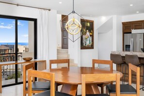 A modern dining area with a round wooden table and chairs, a pendant light, and a view of the sea through large windows.