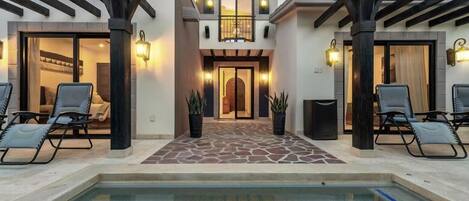 Un área de piscina con sillones de descanso, un patio con suelo de piedra y la entrada de un edificio con puerta y ventanas.