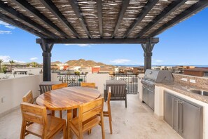 An outdoor patio with a wooden table and chairs, a grill, and a view of the surrounding area.