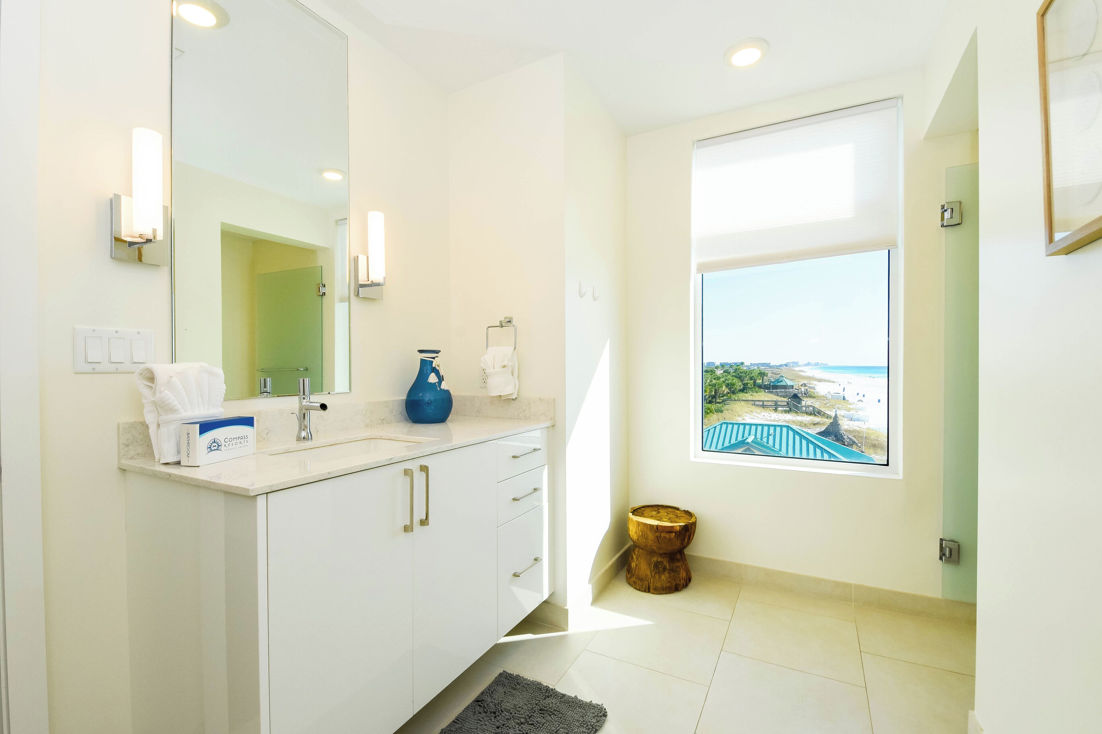 Une salle de bain moderne avec un grand miroir, un meuble-lavabo blanc aux poignées dorées, un vase bleu et une vue sur la plage par la fenêtre.