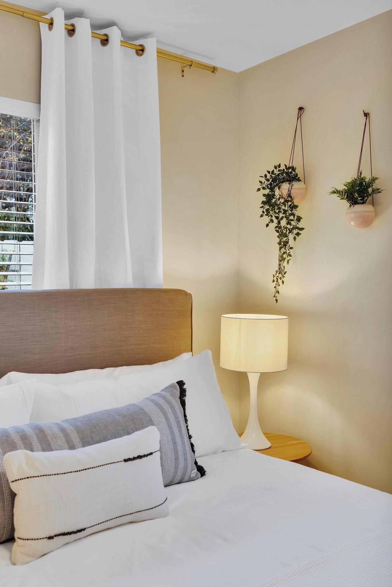 Un lit bien fait, avec une literie blanche et des oreillers à rayures, une lampe de chevet et une fenêtre ornée de rideaux blancs.