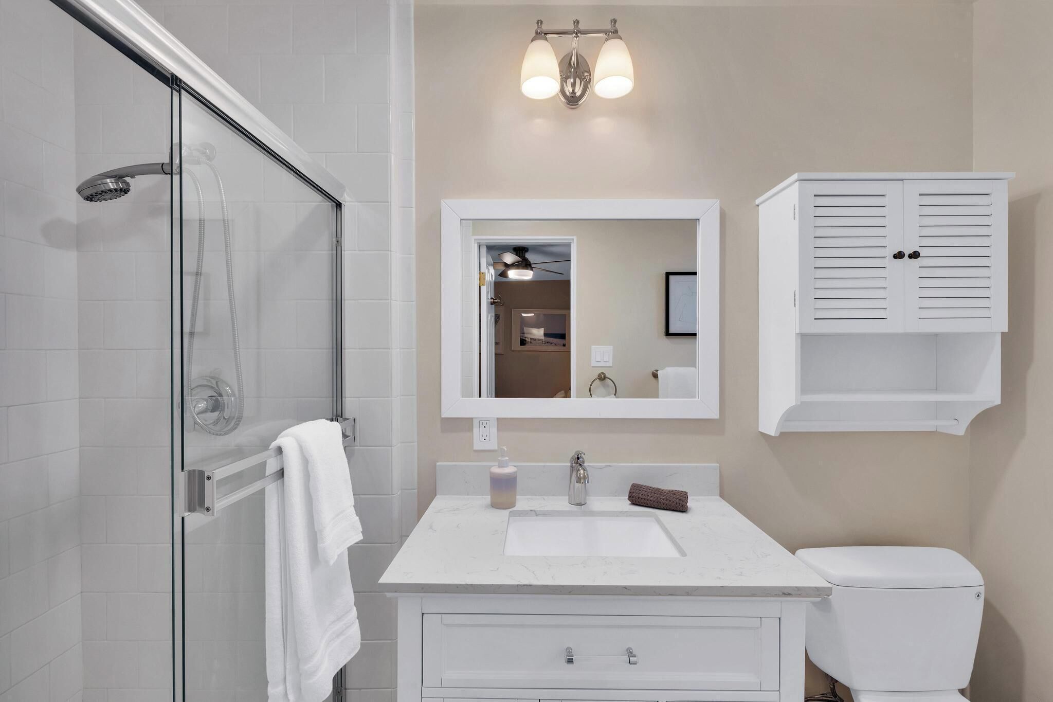 A bathroom with a glass shower enclosure, a white sink with a marble countertop, a mirror above the sink, and a white cabinet with two doors.