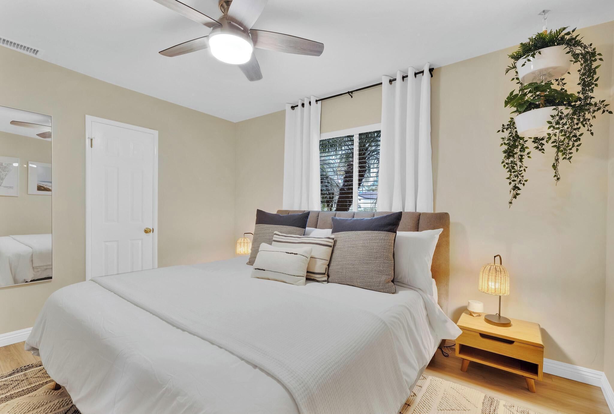 Un dormitorio con cama, ventilador de techo, mesita de noche con lámpara, ventana con cortinas y una planta en la pared.