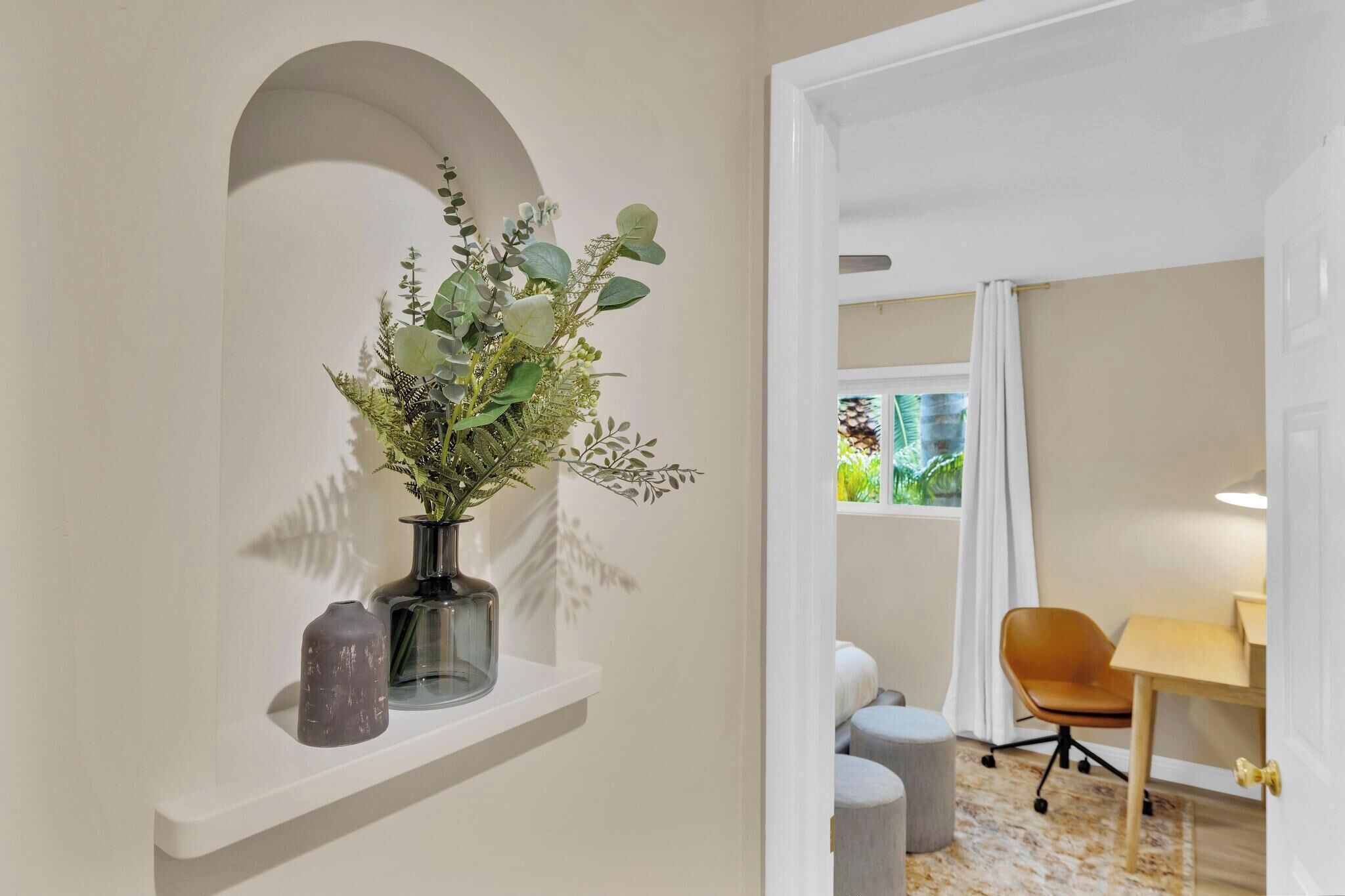 Une pièce moderne avec un mur blanc, une étagère ornée d’un vase de plantes vertes, un bureau avec une chaise et une fenêtre avec des rideaux.