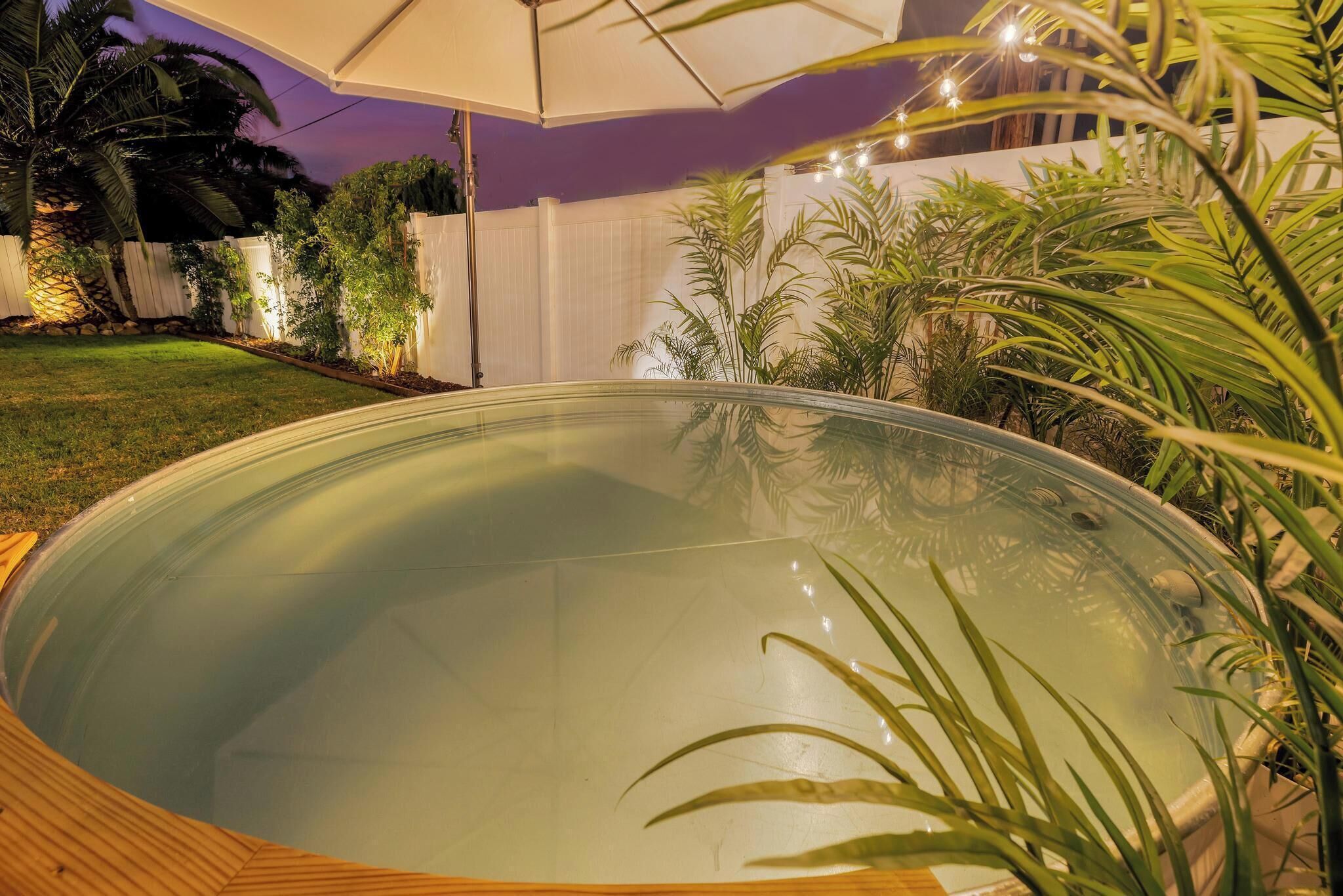 Una piscina con muelle de madera, rodeada de plantas y una valla blanca.