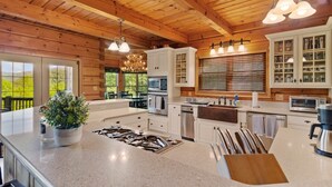 Une cuisine spacieuse dotée d’un grand îlot central, de poutres apparentes en bois au plafond et offrant une vue sur l’extérieur.