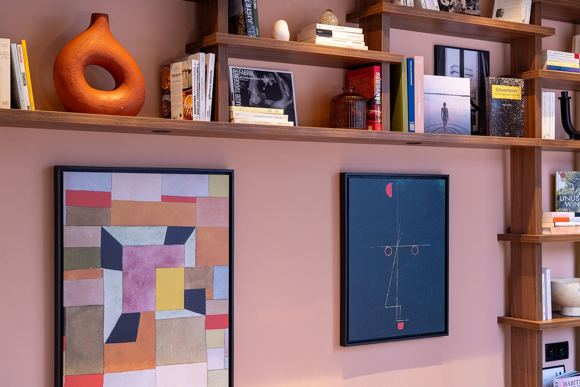 Un mur orné de deux œuvres d’art encadrées et surmonté d’une étagère en bois où sont posés des livres et des objets décoratifs.