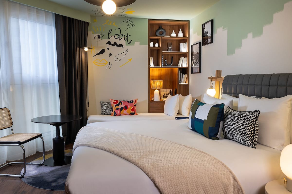 A hotel room with a large bed, a small table, a chair, and a bookshelf.