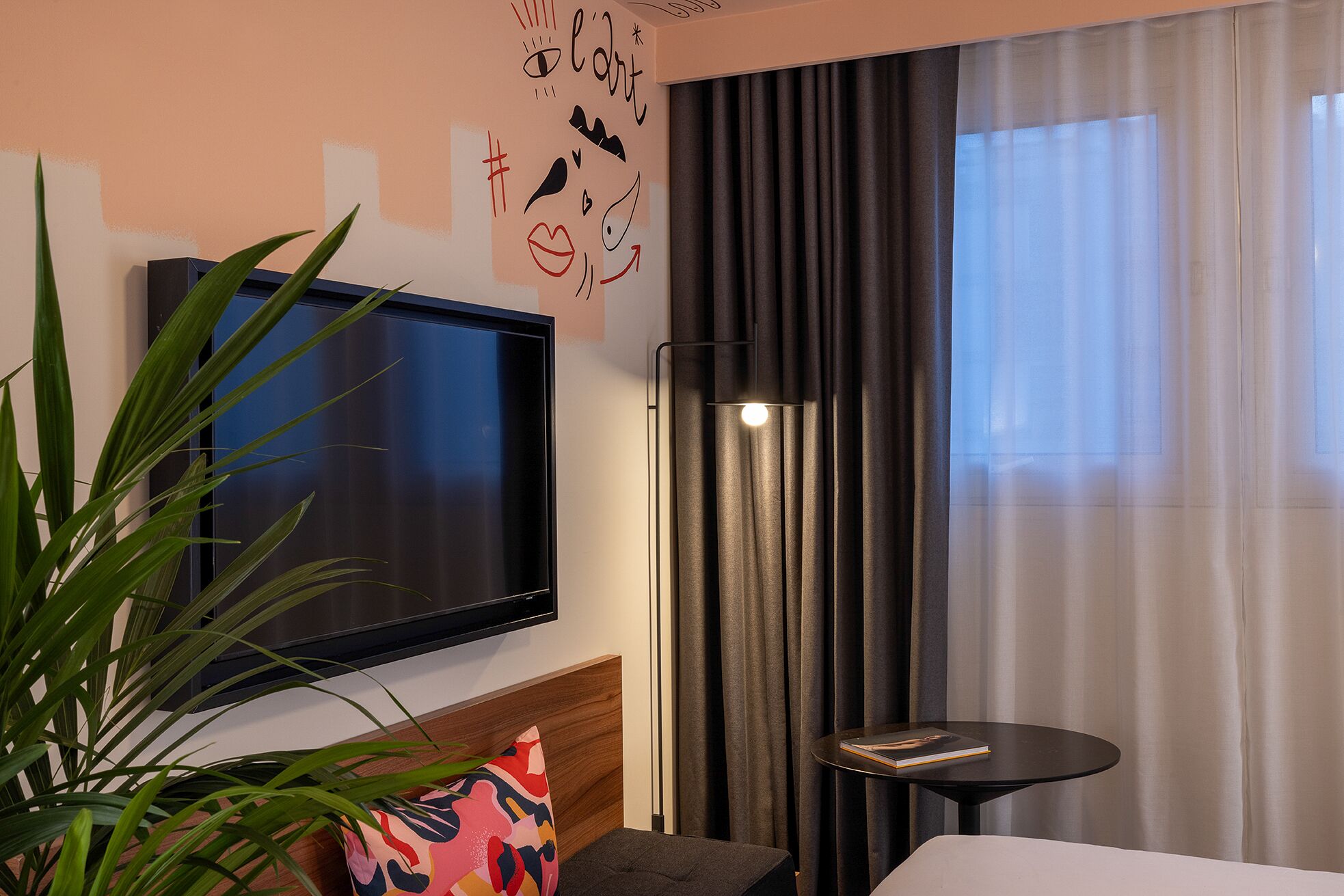 A modern hotel room with a flat-screen TV mounted on the wall, a floor lamp, a small round table, and a potted plant.