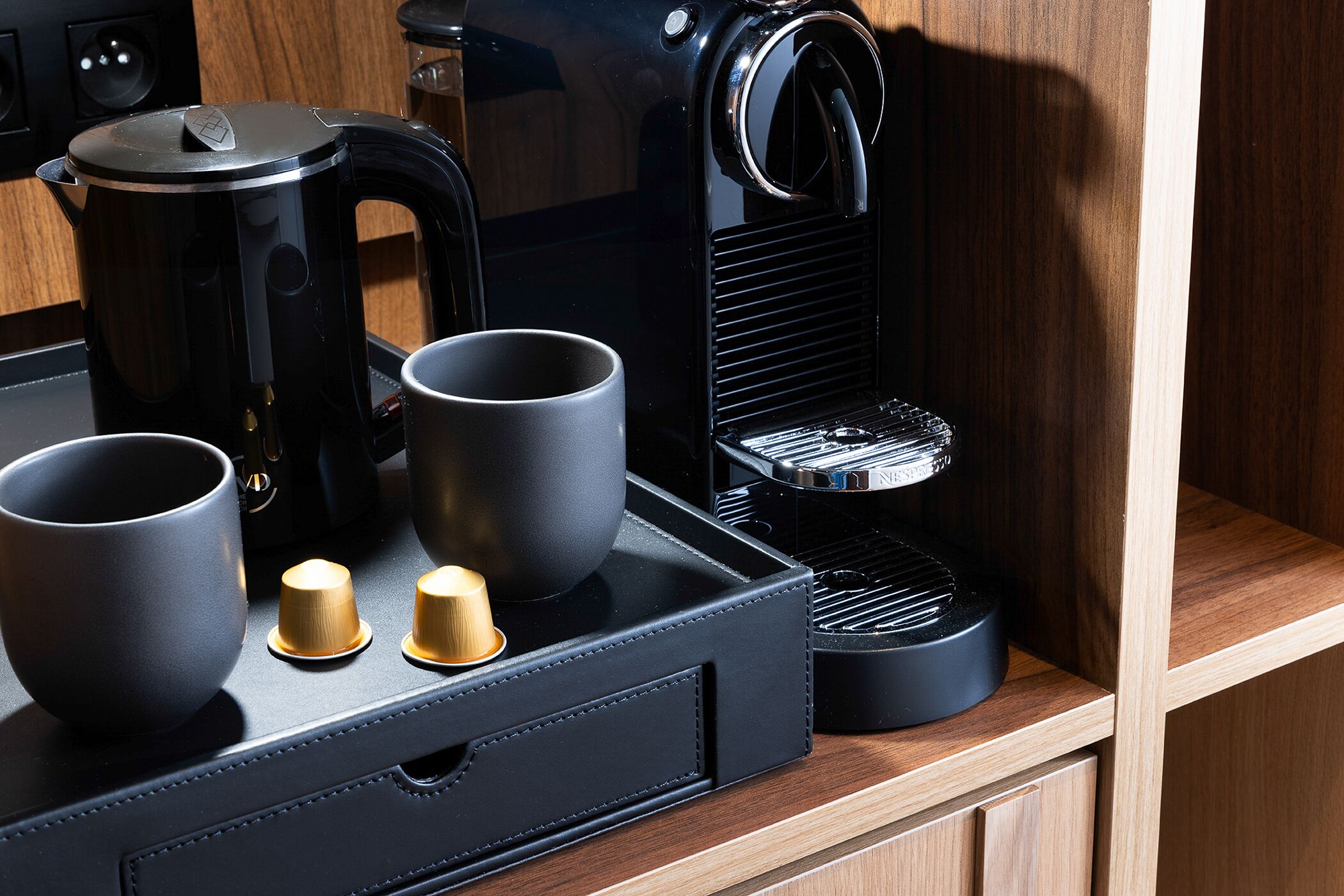 A kitchen area with a coffee maker, two cups, and a kettle on a wooden countertop.