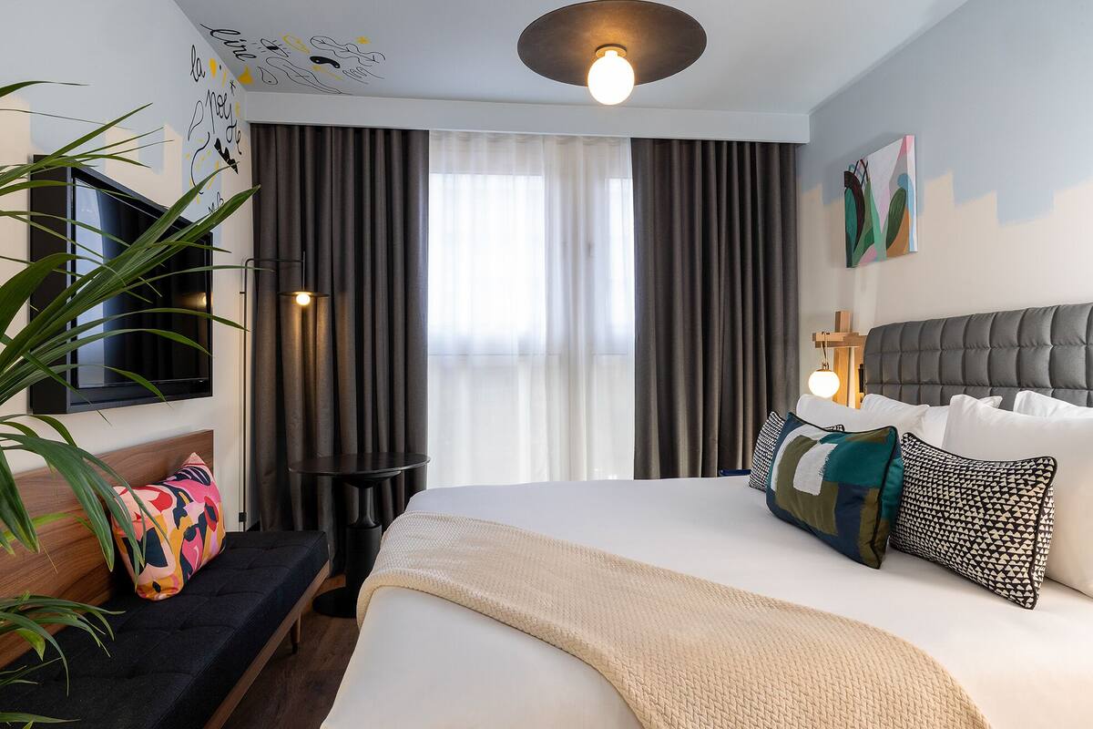 A modern hotel room with a large bed, a TV mounted on the wall, a bedside table with a lamp, and a large window with curtains.