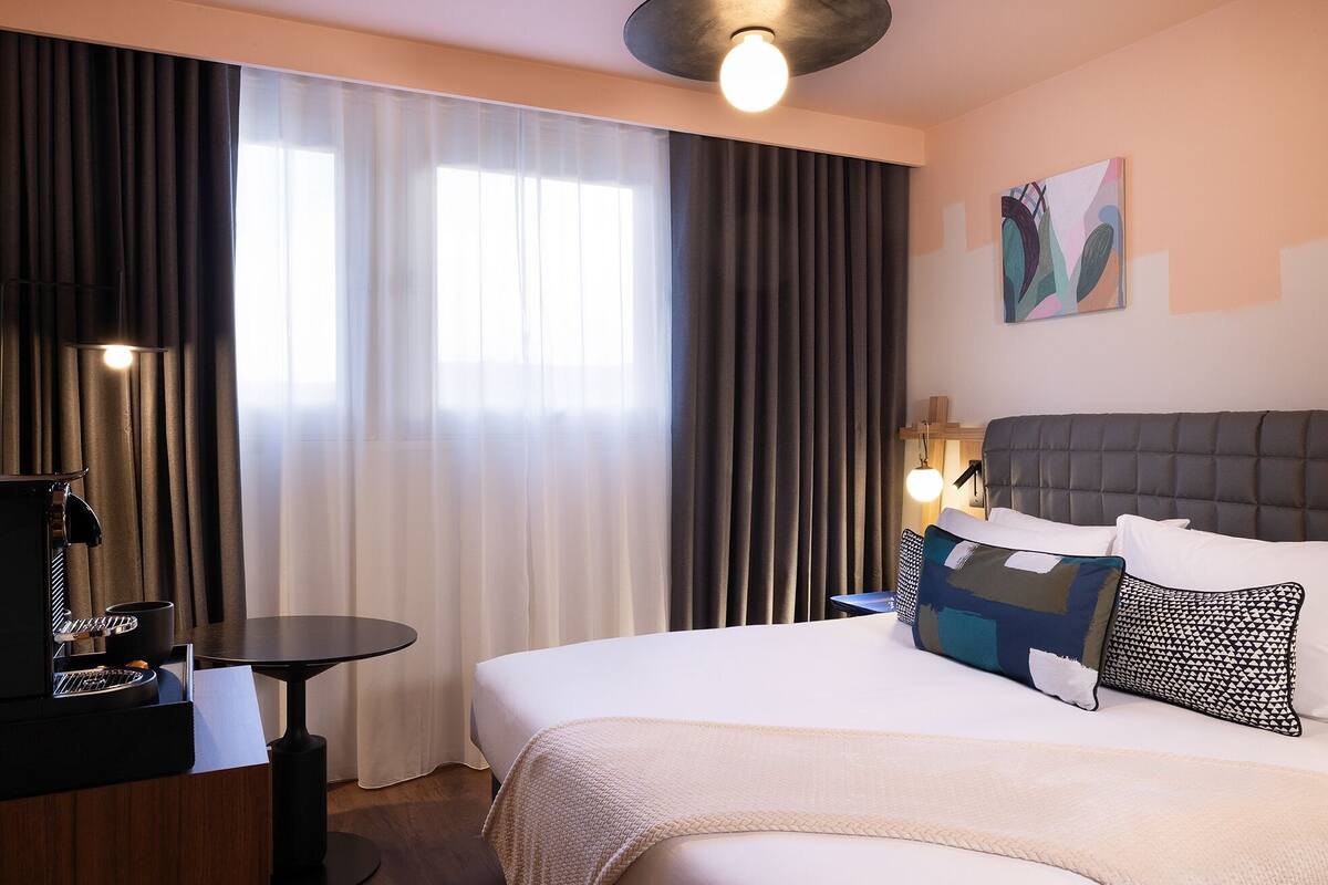 A hotel room with a bed, a desk with a coffee machine, a lamp, and a painting on the wall.