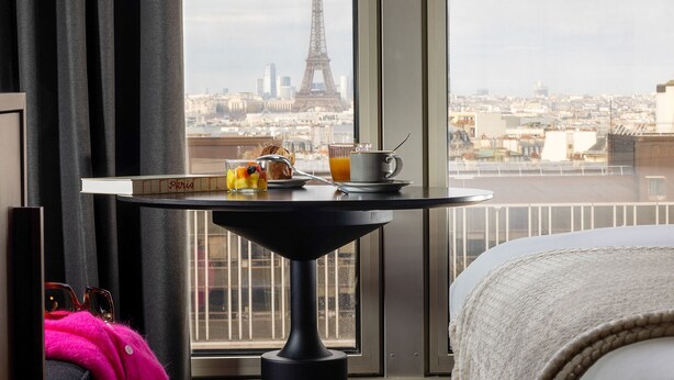 Una habitación de hotel con vista a la Torre Eiffel, una bandeja de desayuno con croissants y zumo de naranja, y una cama con una manta blanca.