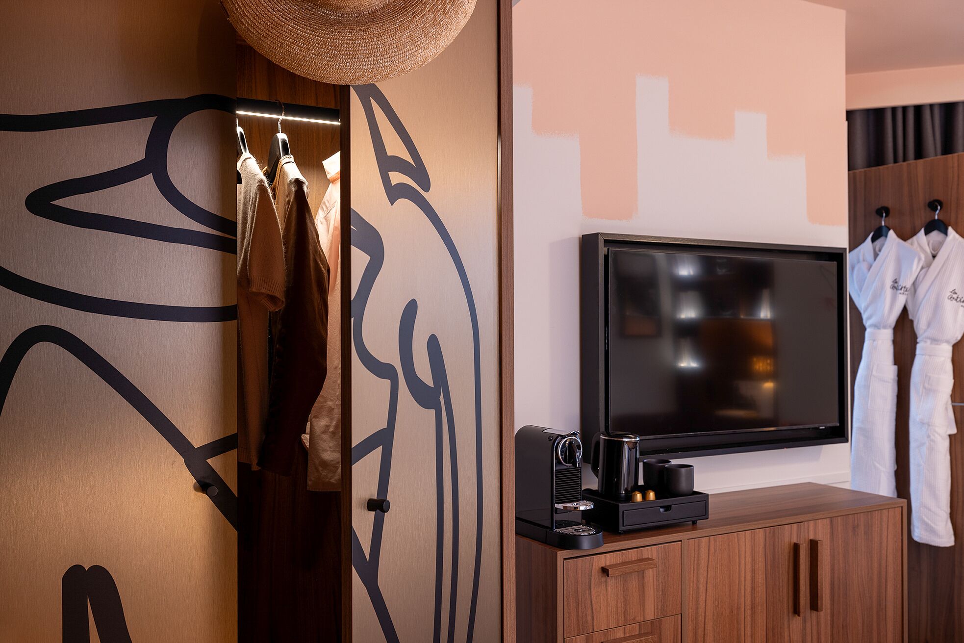 Una moderna camera d'albergo con una grande TV a schermo piatto montata su un mobile in legno, una macchina per il caffè e una parete decorata con arte astratta.