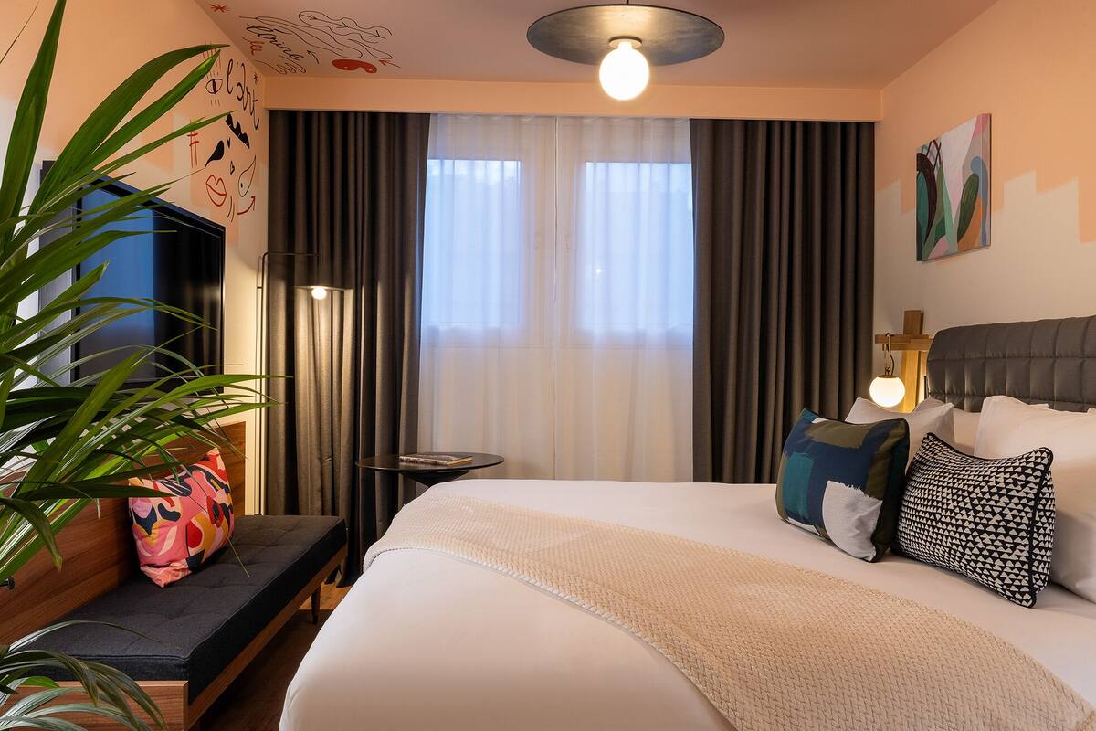 A neatly arranged hotel room with a bed, bedside table, lamp, and a large window with curtains.