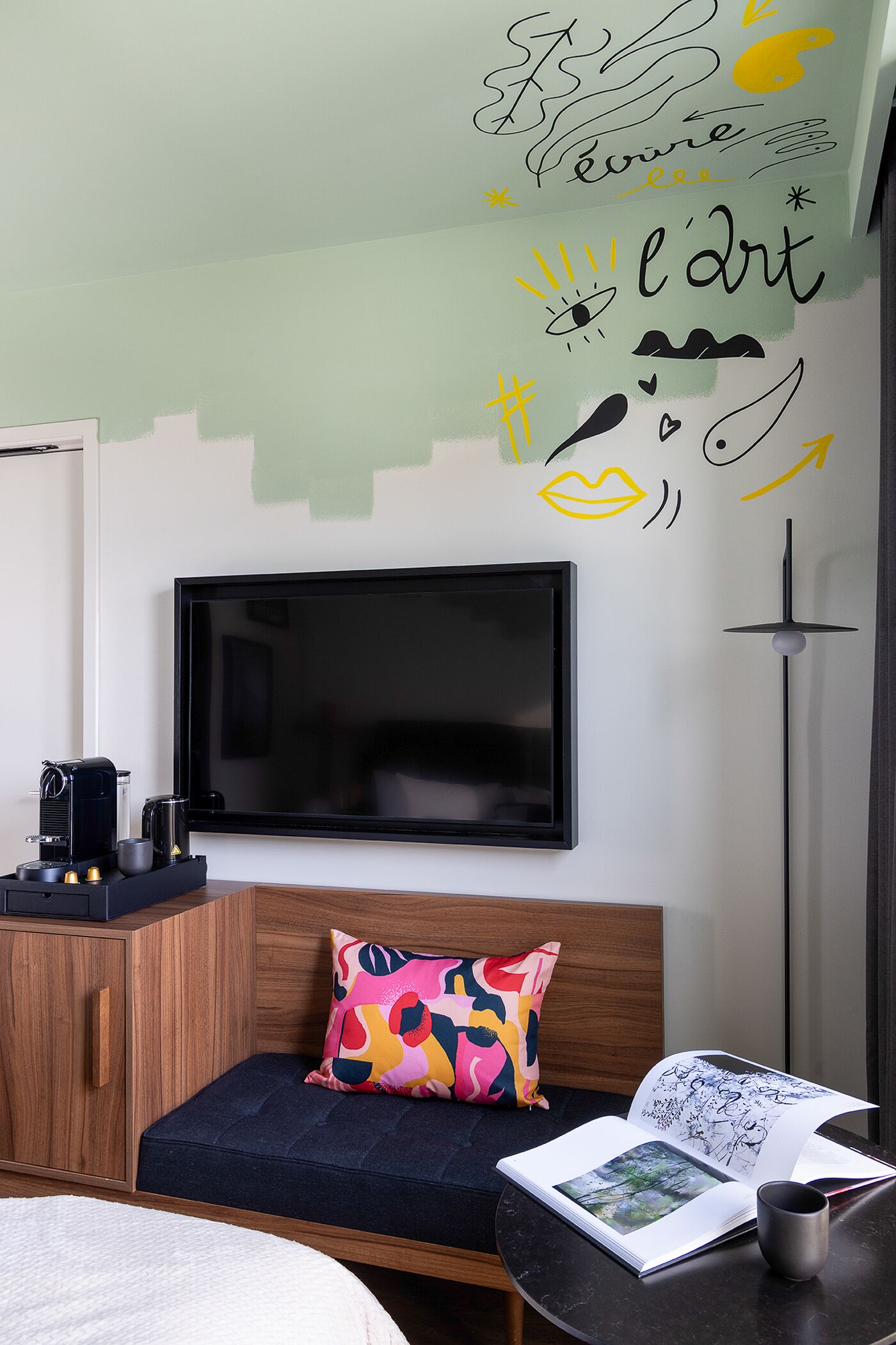 A modern hotel room with a large flat-screen TV mounted on the wall, a wooden cabinet, a sofa with a colorful pillow, a small table with a book and a mug, and a floor lamp.