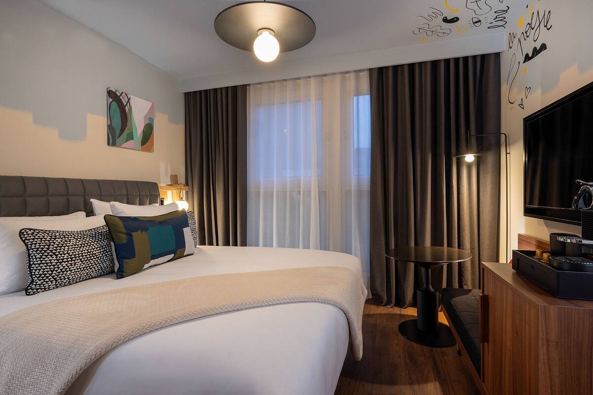 A hotel room with a large bed, a desk, a flat-screen TV, and a view of the city through the window.