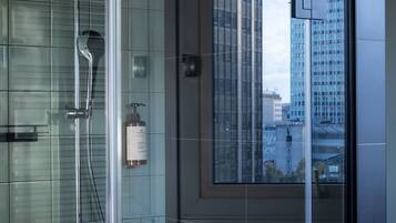 A modern bathroom with a glass shower enclosure, a visible bottle of shampoo, and a view of a cityscape through a large window.